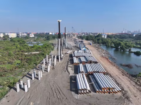 Luftaufnahme einer Baustelle mit gestapelten und aufgestellten Betonpfählen, zwei Rammgeräten und einem Kran, daneben Grünflächen und ein Gewässer